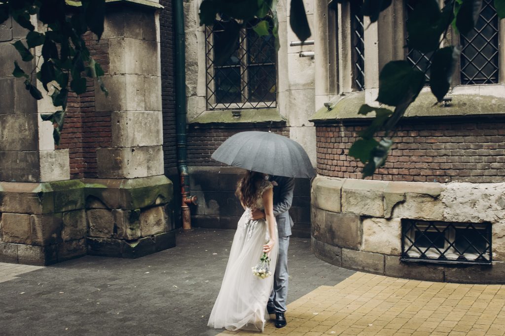 Ein sich im Regen unter einem Regenschirm küssendes Brautpaar steht vor einer alten Kirche